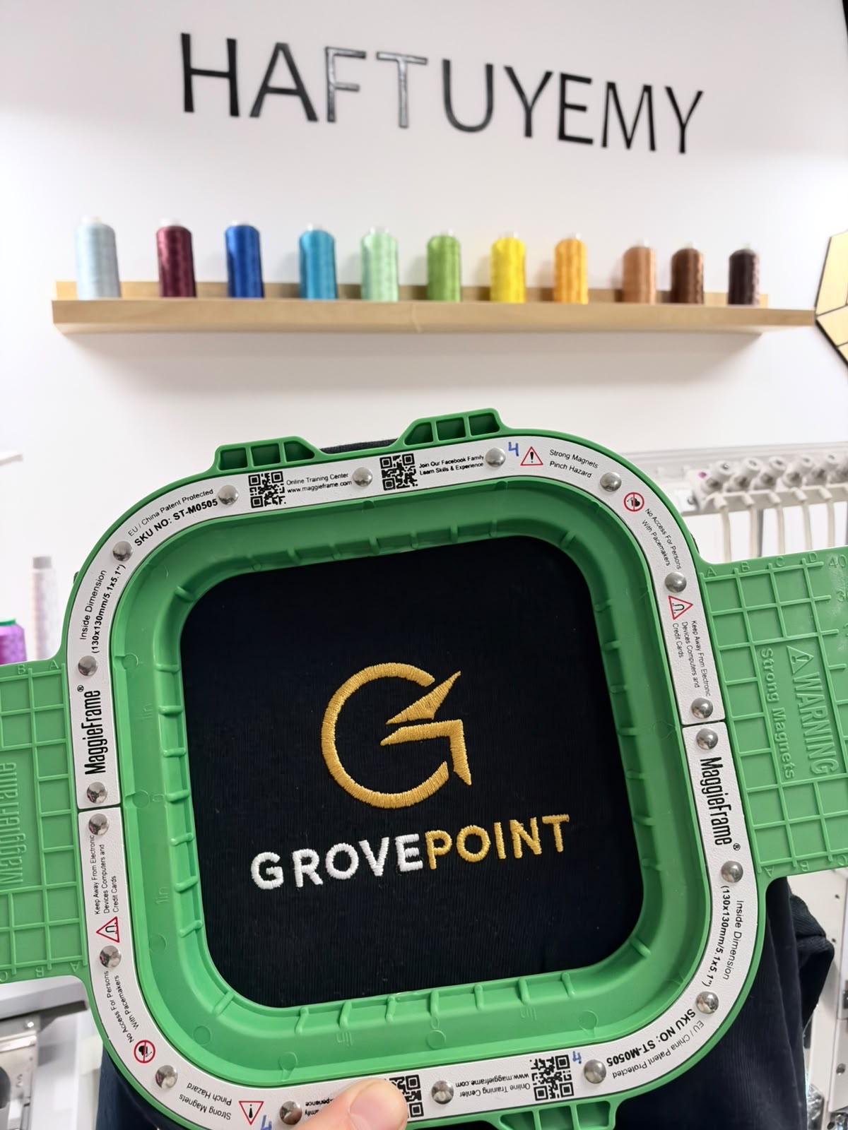 Koszulka z wyhaftowanym logo GrovePoint w ramie haftującej, w tle pracownia Haftuyemy